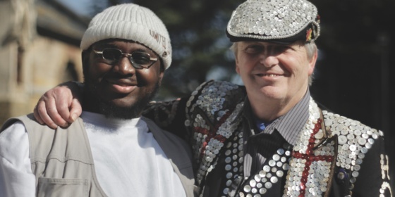 A pearly king puts his arm around a man in a beanie hat