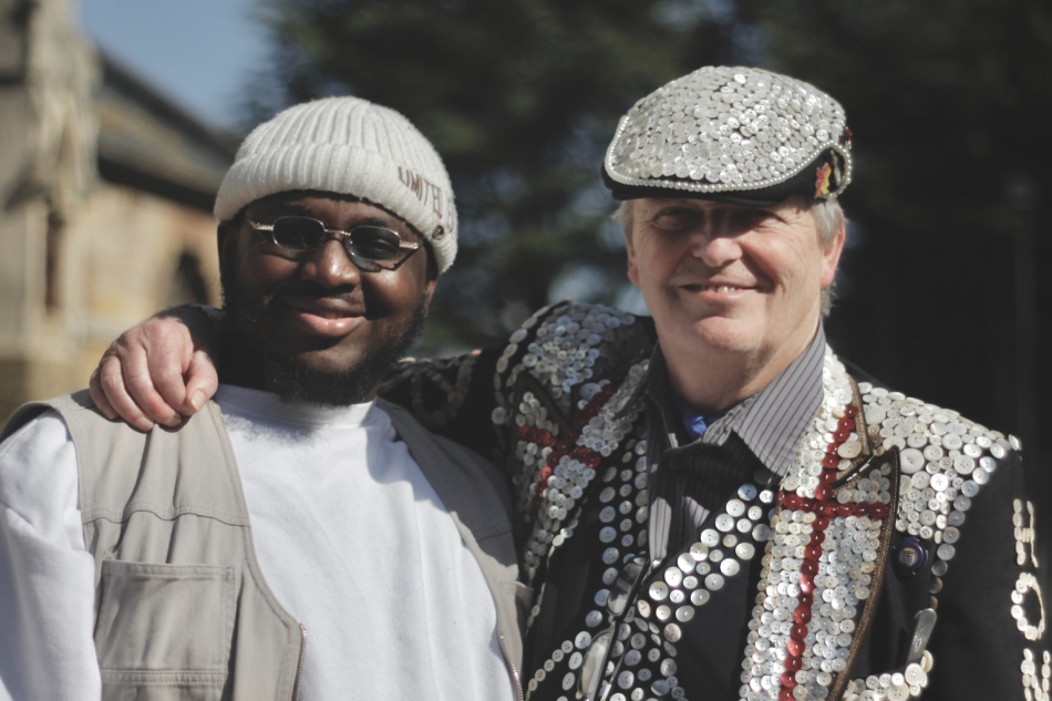 A pearly king puts his arm around a man in a beanie hat