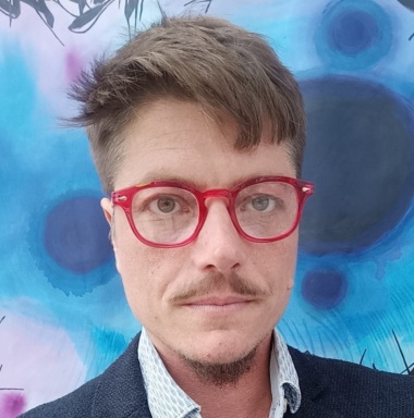 White man with red glasses in front of painting in shades of blue, black, purple and white.