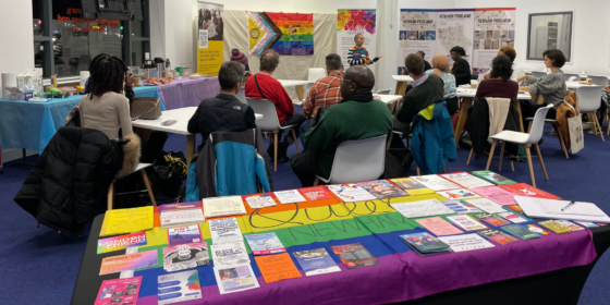 A group of people listening to a Queer Newham facilitator. before a table covered in a Pride flag with leaflets on it.