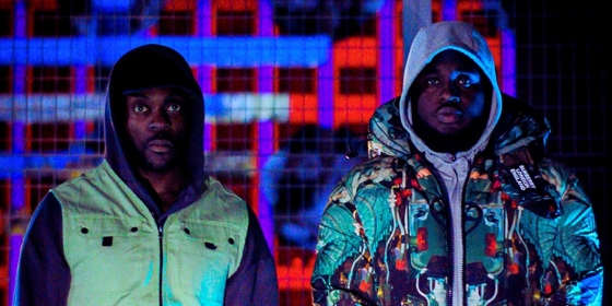Prince & Ebs of Ruff Sqwad Arts Foundation stadning in front of a brightly-lit Millenium Mills at night on the Royal Docks in Newham