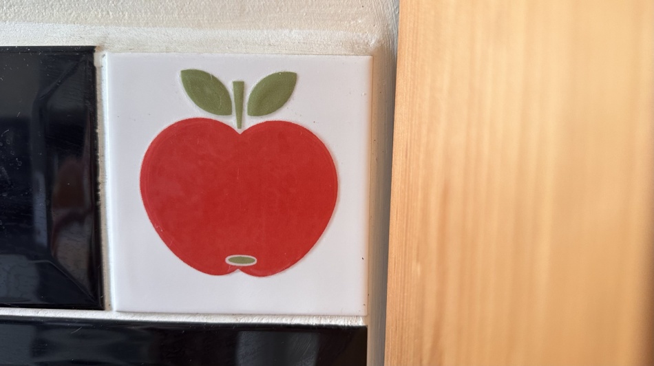 A decorative kitchen tile featuring a red apple, surrounded by wood shelving on the right and black tiles on the left
