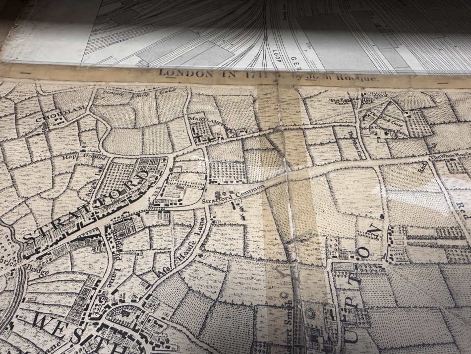 photograph of a pile of maps from a visit to Stratford archives. On top is one that shows roads and a few buildings at Stratford, then fields. The map is damaged and held together with sticky tape at the centre which looks darker. This fault-line is where the estate is now.